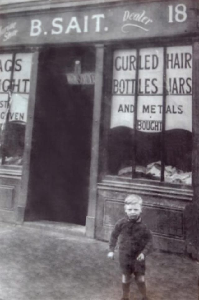 Black and white photograph of the original B. Sait shopfront, its windows painted with signs for rags, curled hair, bottles, jars and metals bought, and a young boy standing out front